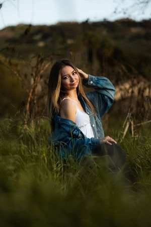 Young woman in denim jacket and white dress sitting in tall green grass outdoors during golden hour.