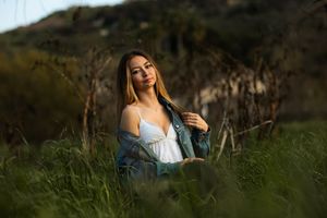 Young woman in white dress and denim jacket sitting in tall green grass with golden sunlight in nature.