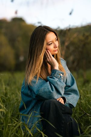 Young woman in oversized denim jacket sitting in green grass, looking down in soft natural light.