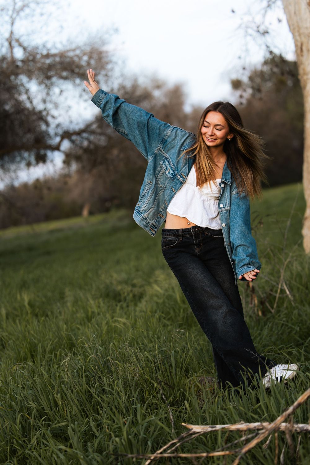 Young woman in oversized denim jacket, white crop top, and black jeans poses playfully in a lush green field.