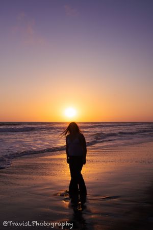 Silhouetted figure kneeling on beach during vibrant orange sunset.