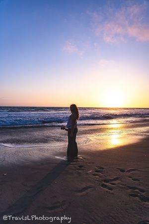 Silhouetted figure walking along beach shoreline at sunset with footprints in sand.