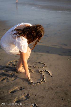 Person in white dress drawing heart shape in beach sand during golden hour.