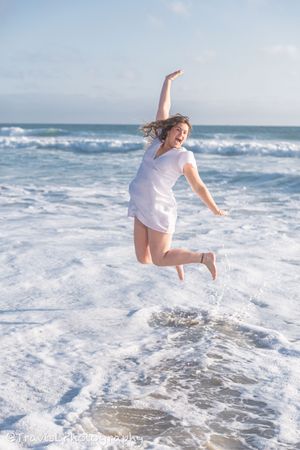 Person in white dress jumping joyfully through ocean waves at the beach.