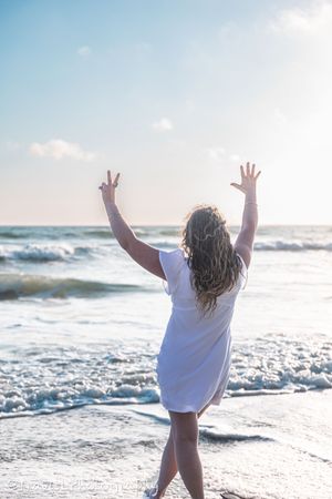 Person in white dress celebrating with arms raised at beach during sunset.