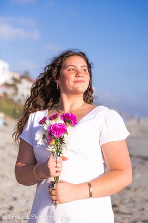 Person in white holding pink flowers on sunny beach with blue sky background.