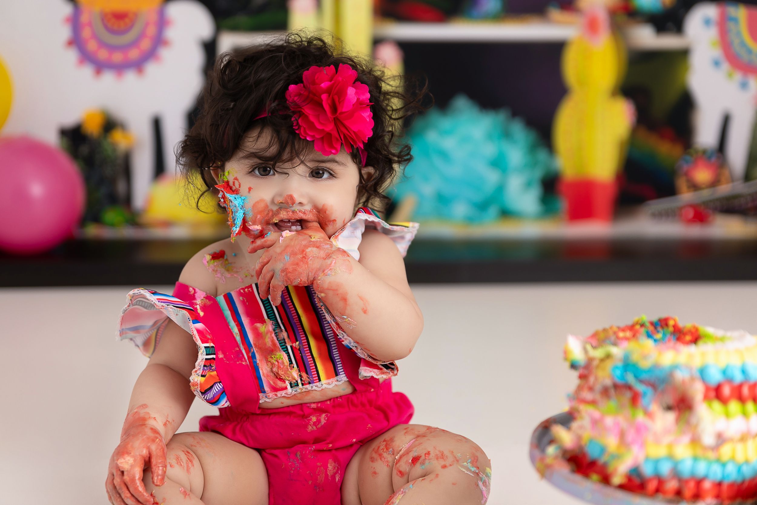 A baby in a bright pink outfit sits with a colorful cake during a festive first birthday celebration party.