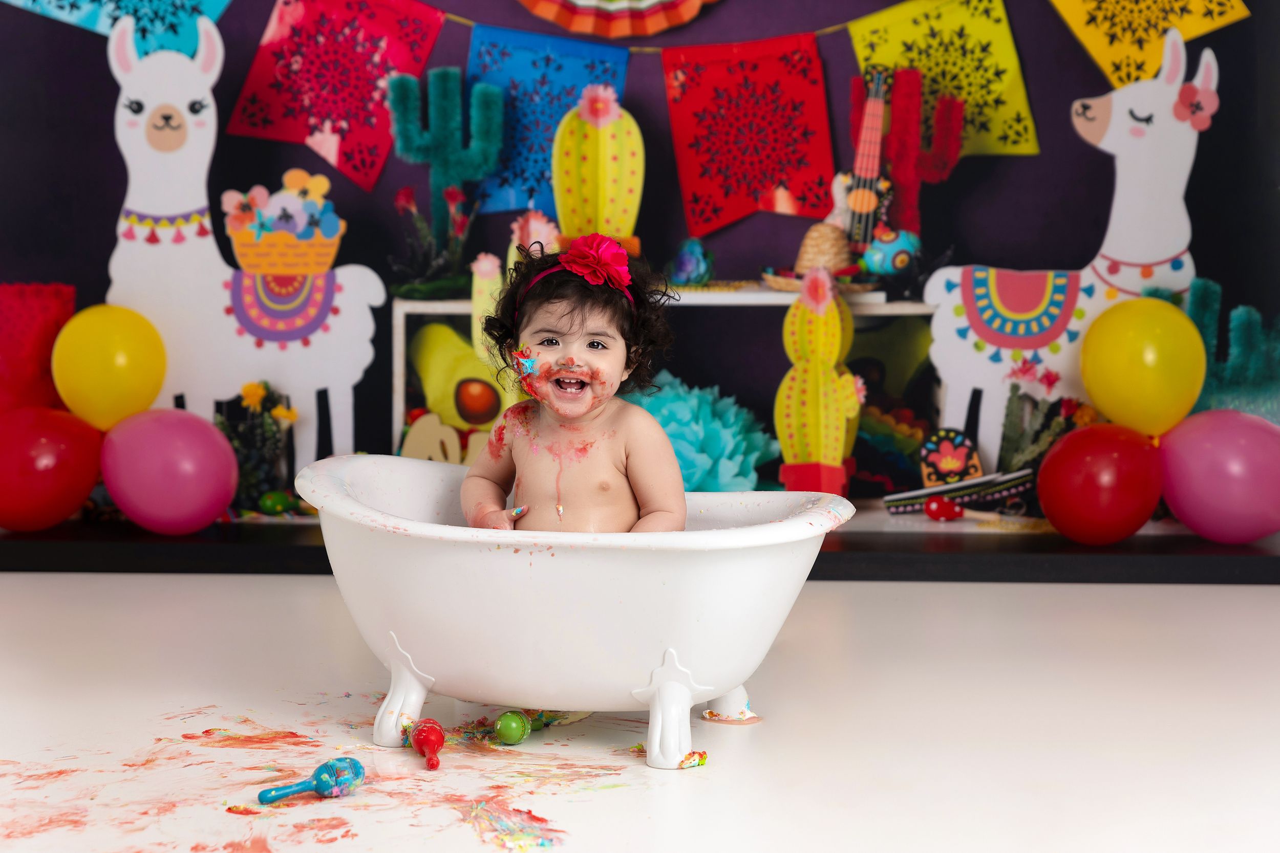 Colorful fiesta-themed party setup with bathtub, llama decorations, balloons and festive banners against dark backdrop.