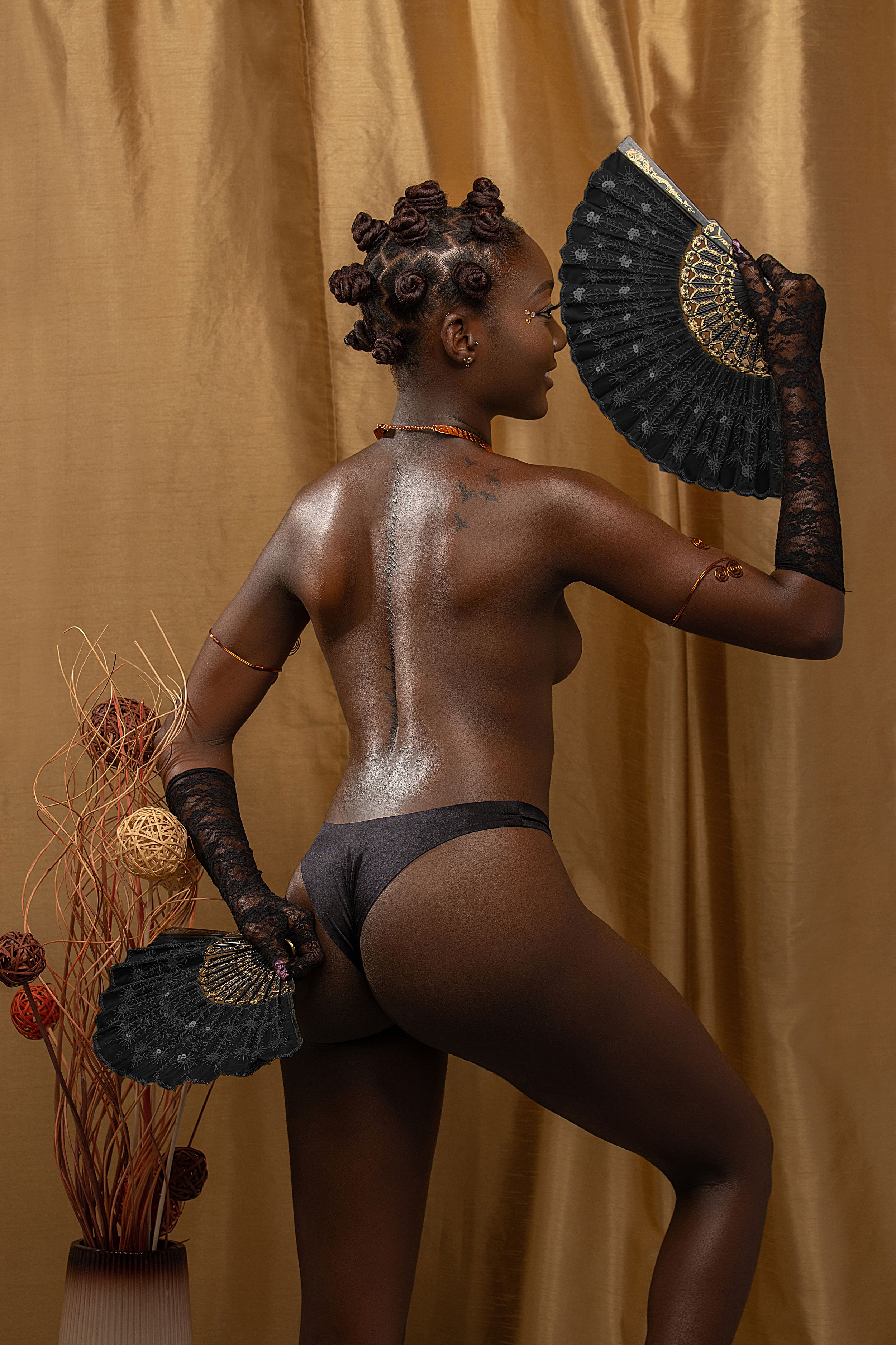 A woman poses with decorative fans wearing black lingerie against a golden draped backdrop.