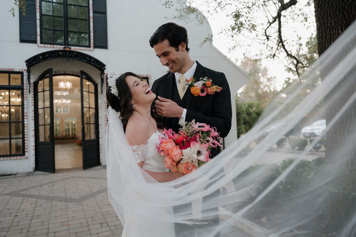 A bride in a white dress holds colorful coral flowers while her veil flows in the breeze during an outdoor wedding ceremony.