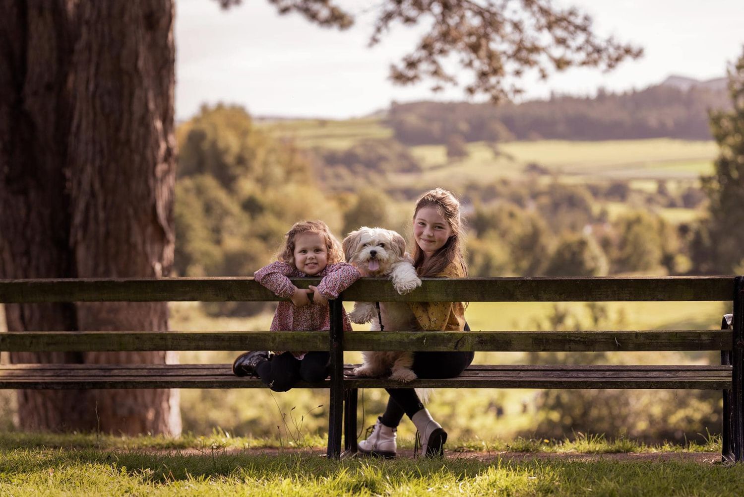 Two children and a small dog sit together on a rustic wooden bench in a pastoral countryside setting.