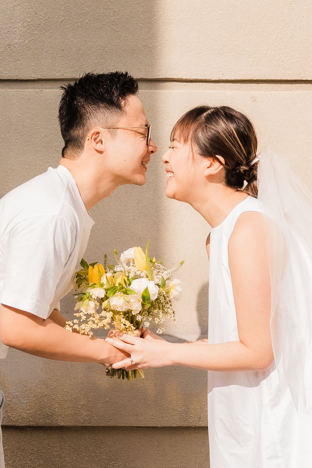A romantic couple in white outfits share a tender moment while holding a yellow and white flower bouquet.