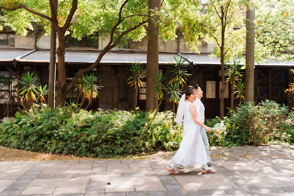 A couple strolls along a garden path surrounded by lush greenery and trees.