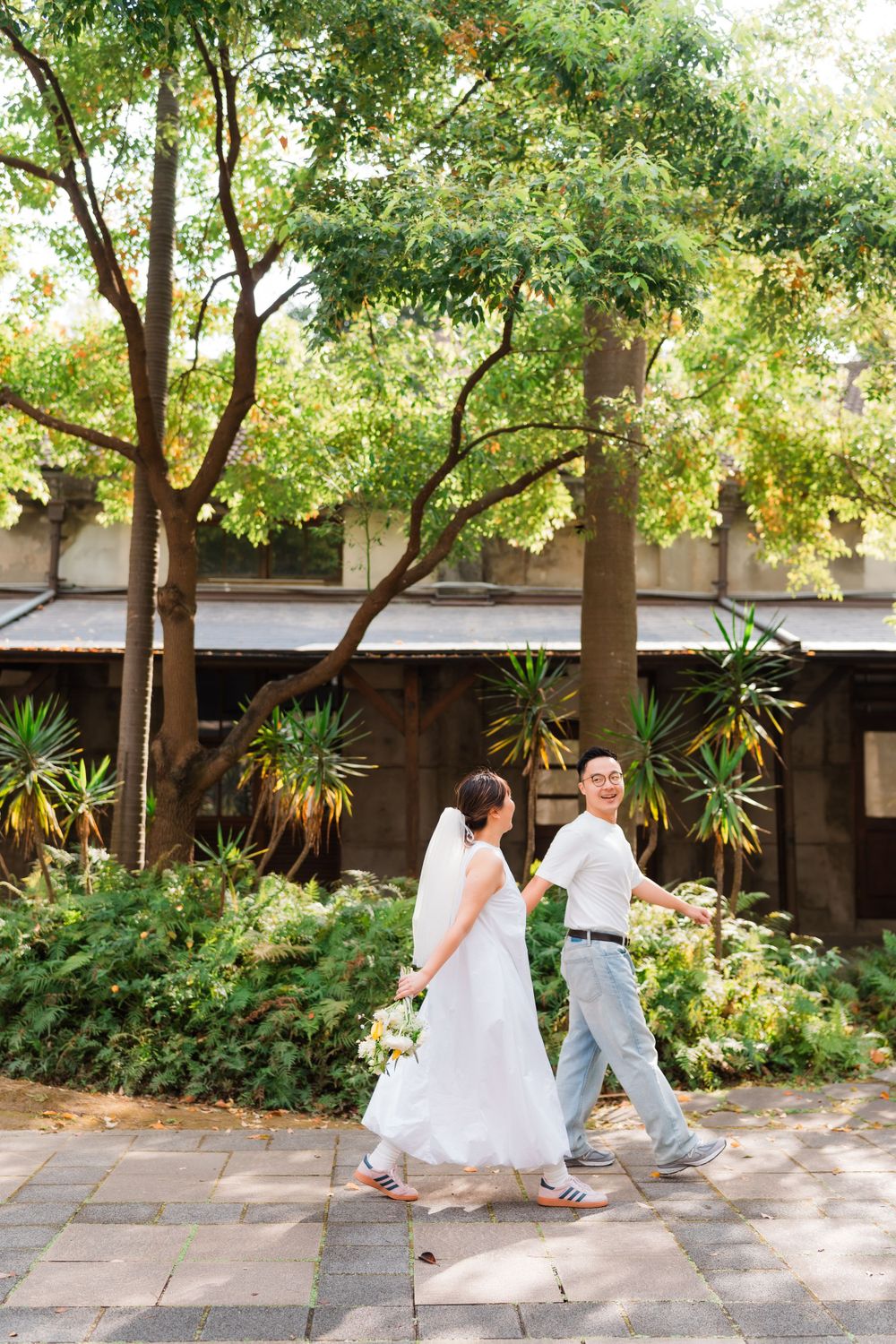 A couple in white walking together along a tree-lined pathway with tropical plants.