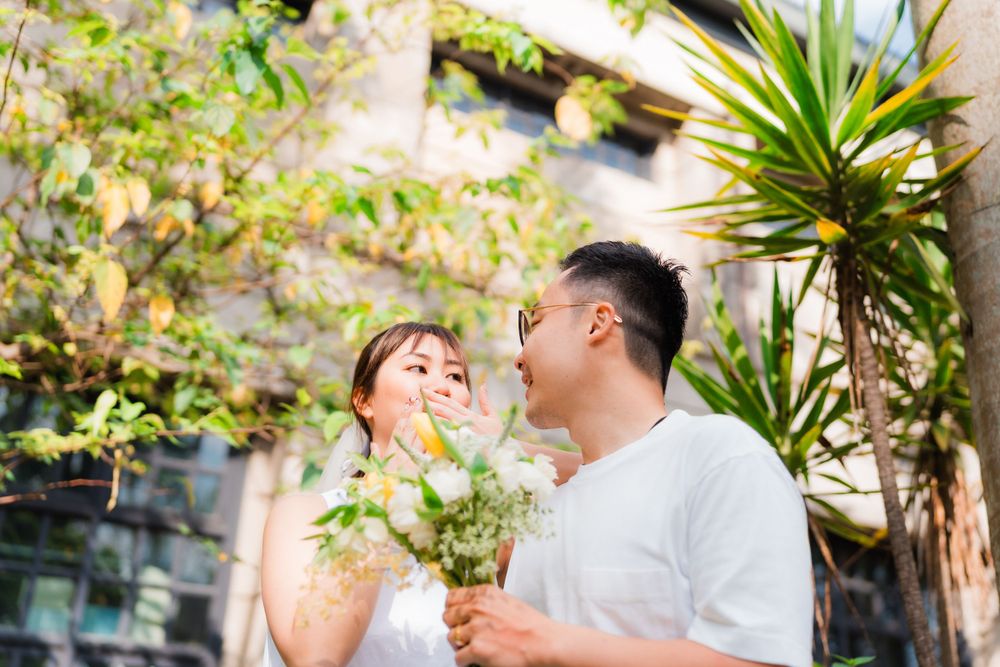A joyful couple in white outfits share a laugh while holding a bouquet outdoors.