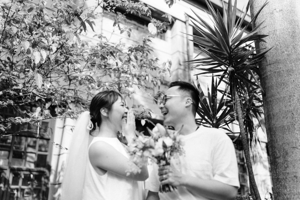 Black and white photo of couple in white sharing laughs while holding flowers outdoors.