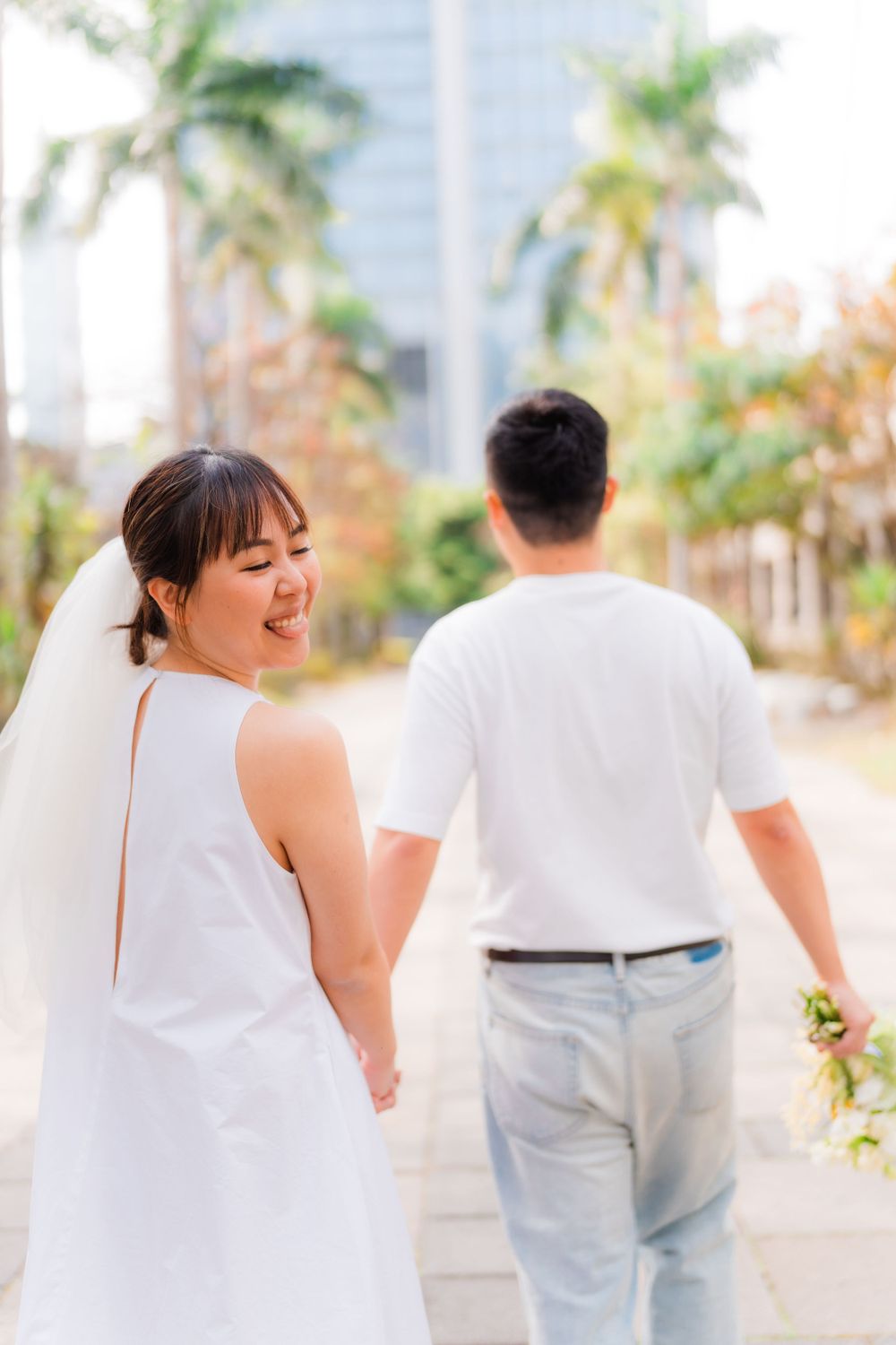 Couple in white walks along palm-lined path with bride looking back at camera.