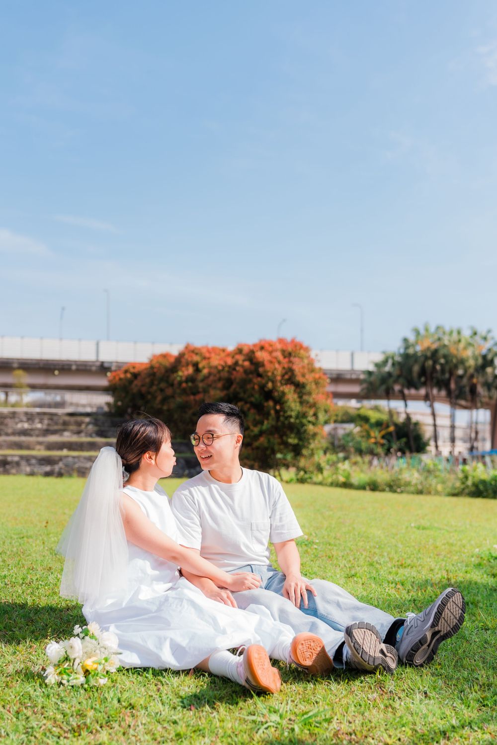 Couple in casual white attire sits together on grass with orange flowering tree in background.