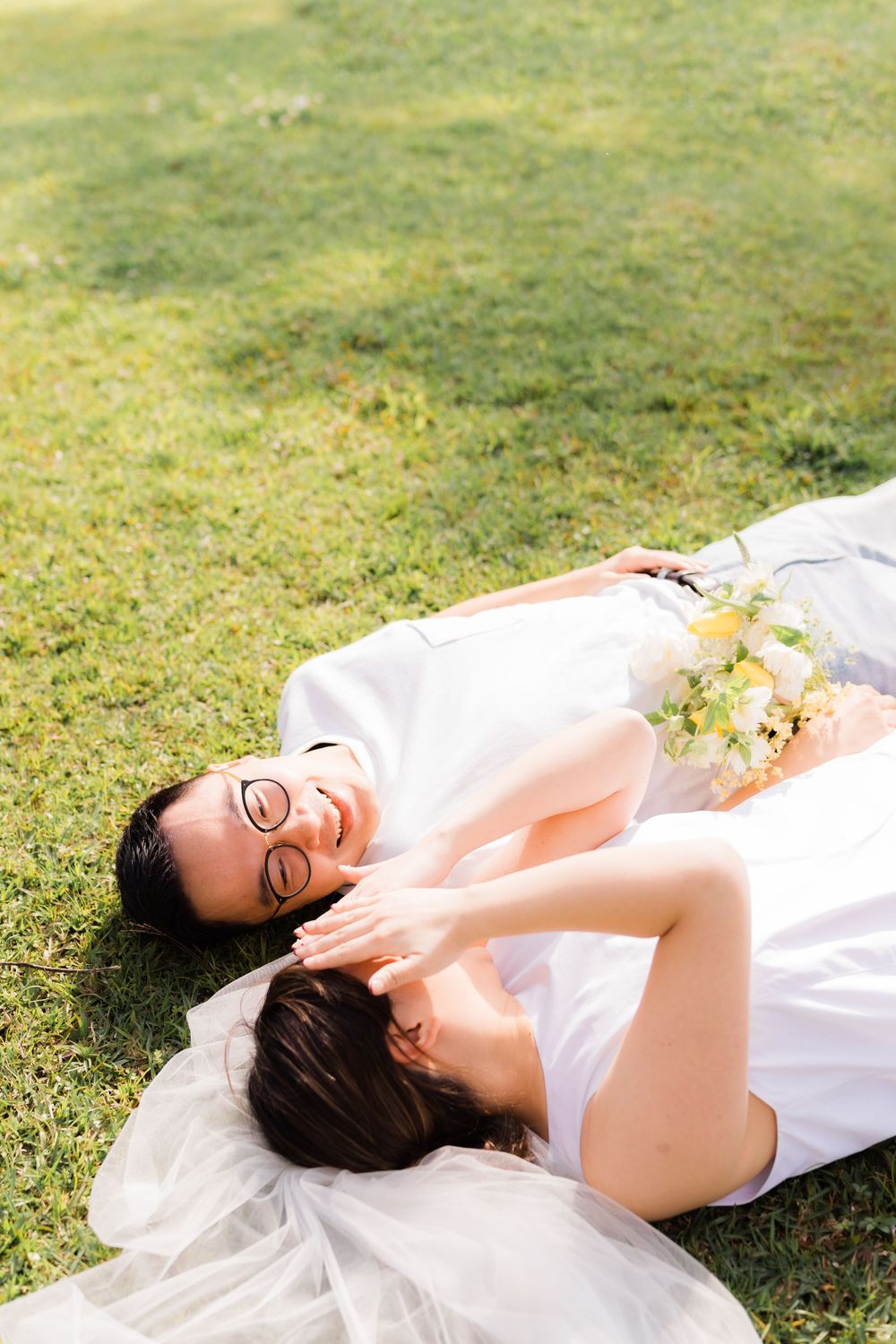 A couple lying on grass in white outfits sharing a peaceful moment with flowers nearby.