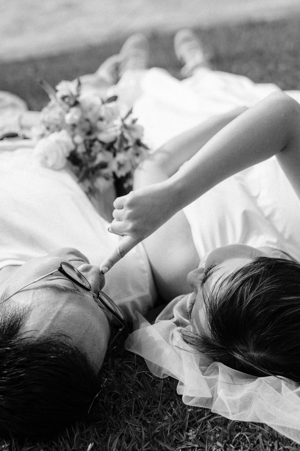 A black and white image of a couple resting on grass with flowers beside them.