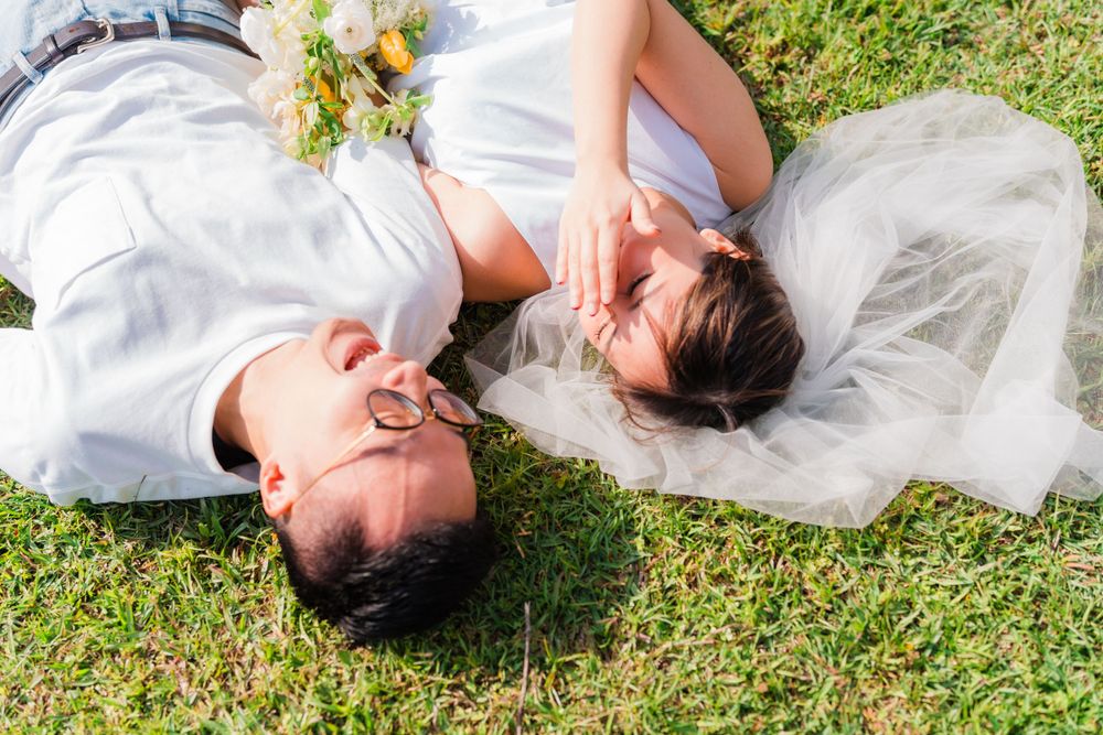 A couple laughs while lying on grass together in matching white outfits.