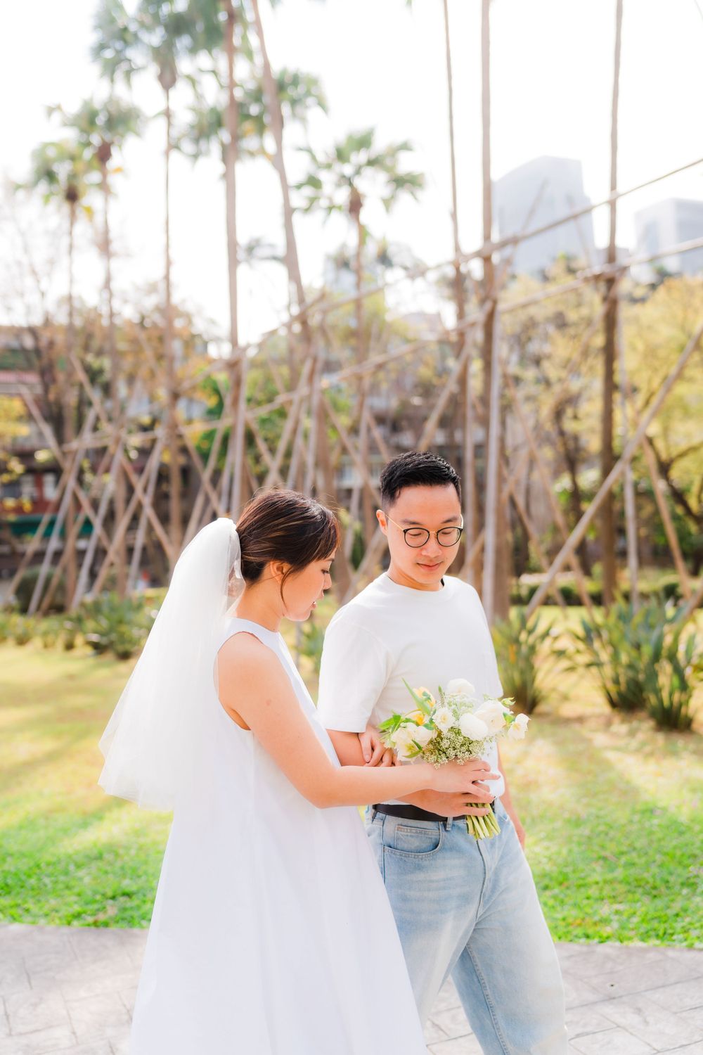 Couple in white casual attire examines bouquet together in tropical garden setting.