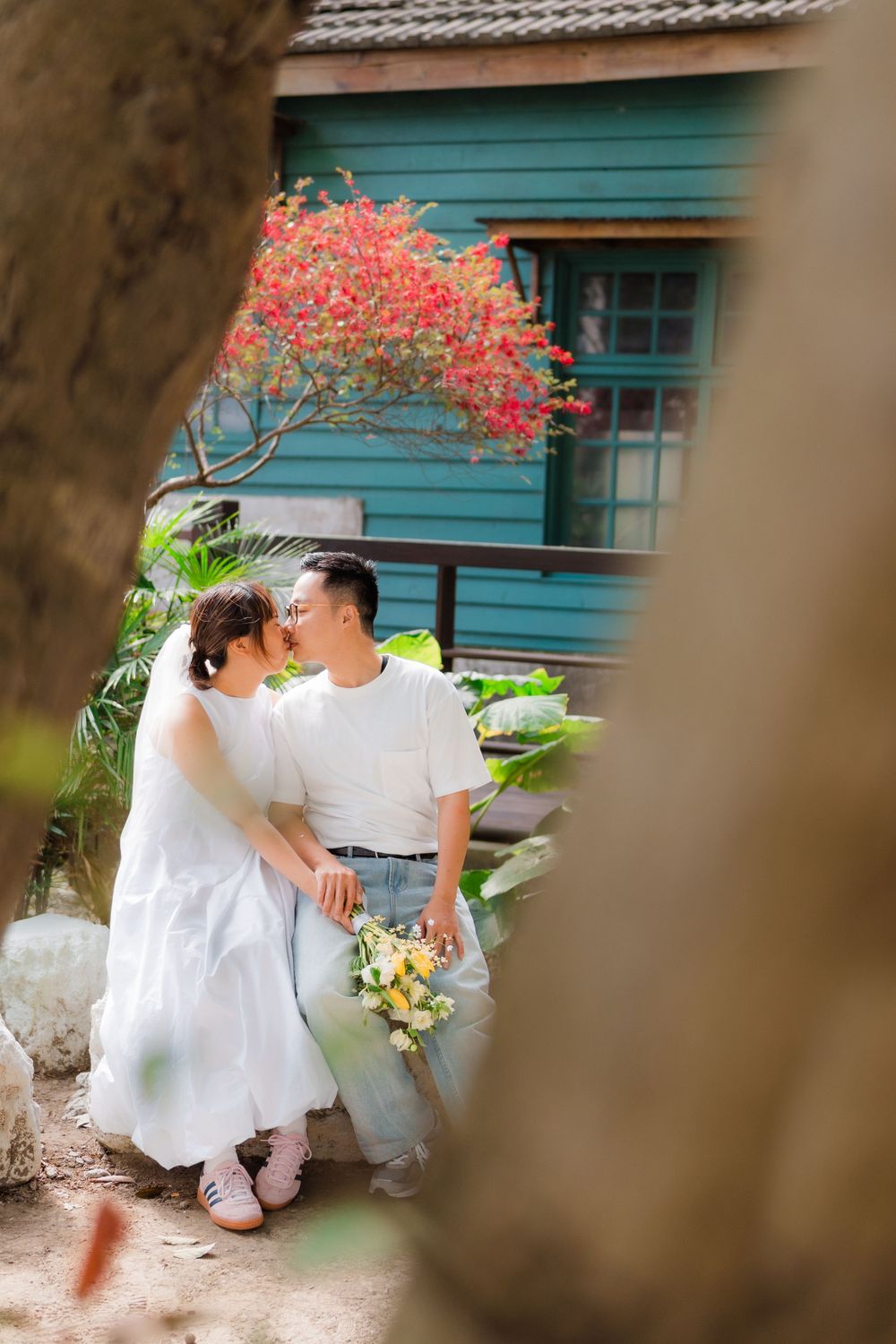 A romantic couple embraces in front of a turquoise building with blooming coral flowers in the background.