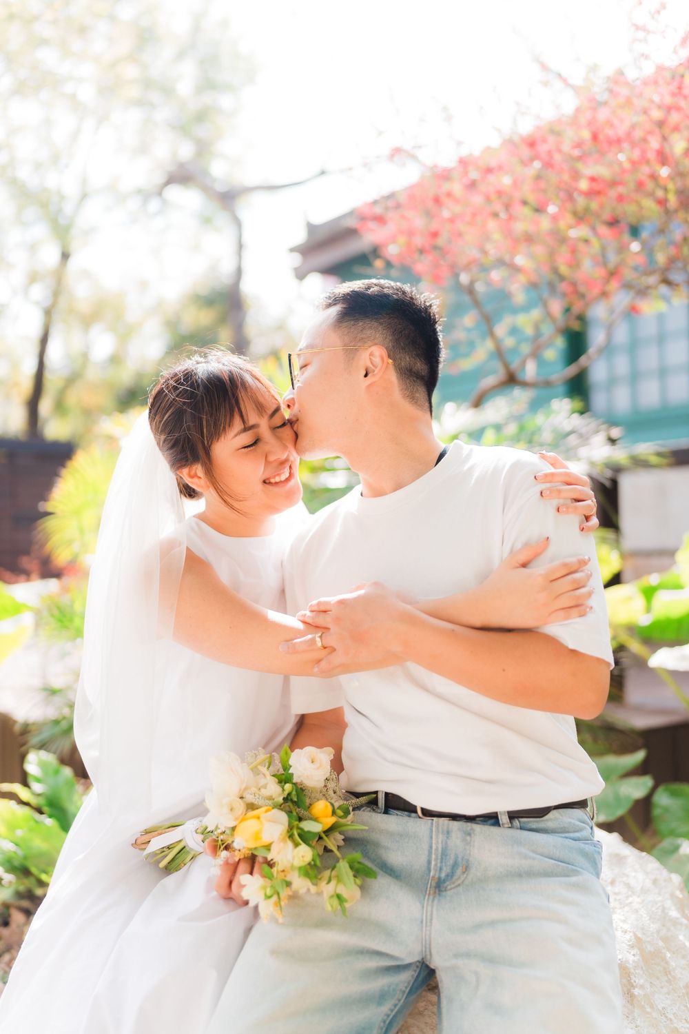 Happy couple in white embraces while holding floral bouquet in autumn garden setting.