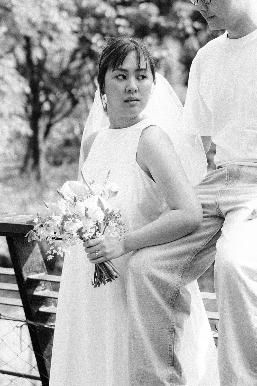 A bride in a white dress holds a bouquet while sitting on a park bench.