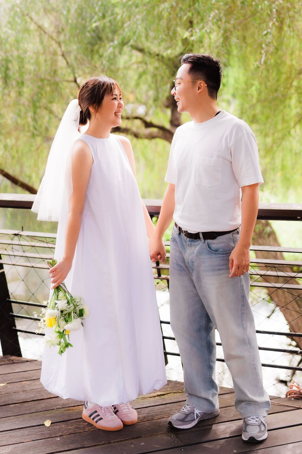 Couple in casual white attire share a happy moment on wooden deck with willow trees in background.