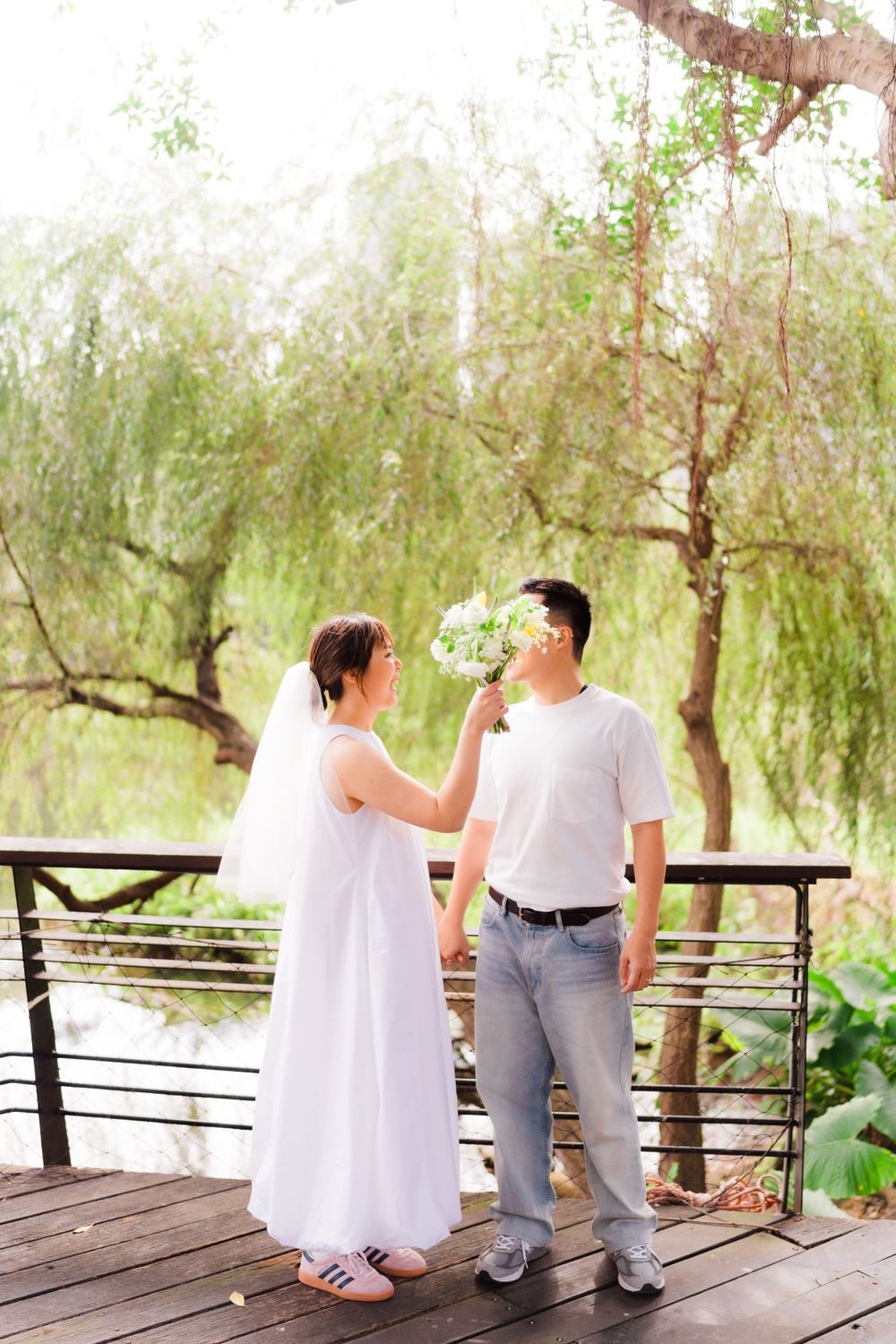 A couple shares a playful moment on a wooden deck surrounded by willow trees.