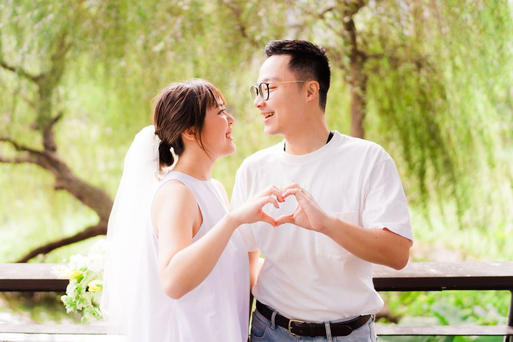 Couple in white makes heart shape with hands while standing on outdoor deck.