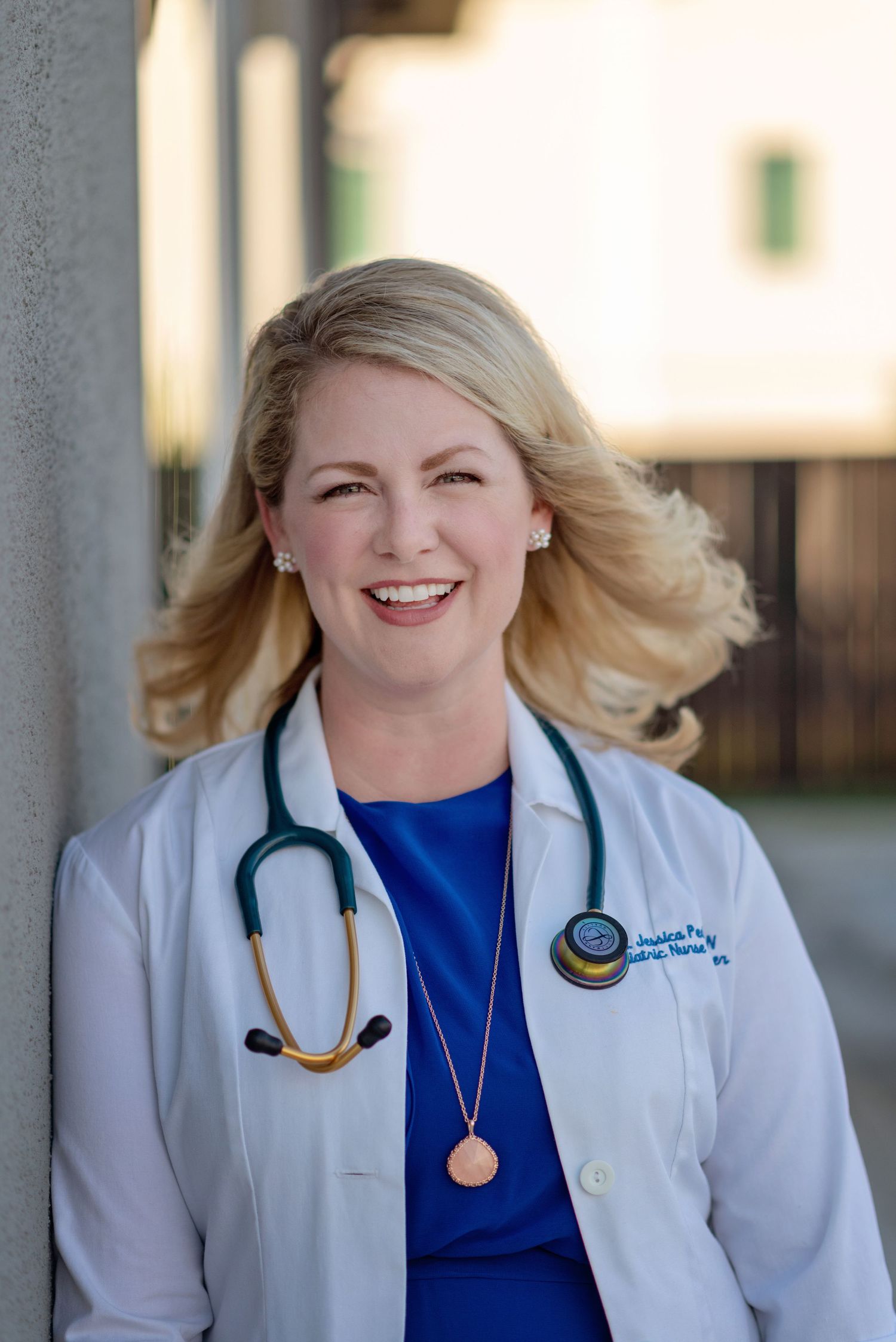 headshot of smiling nurse practitioner leaning against wall wearing white coat and stethoscope
