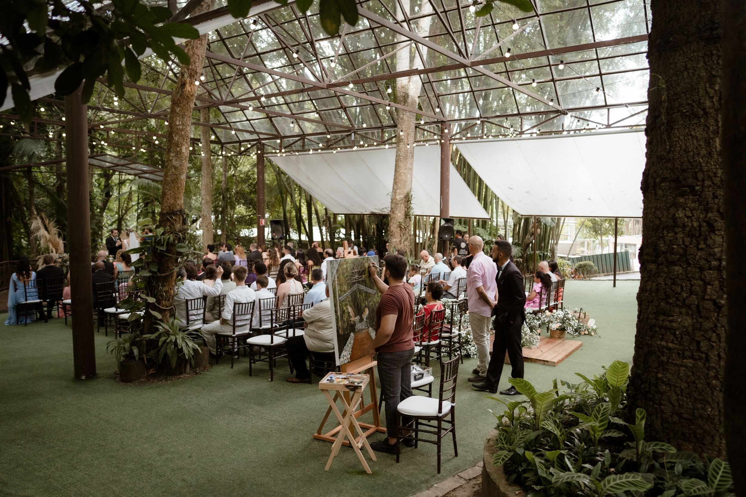 Wedding guests gather under a covered outdoor reception area decorated with string lights and greenery.
