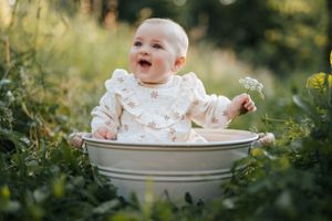 Una bimba seduta in una tinozza circondata dal verde di un prato, tiene in mano un fiore e sorride alla mamma che la sta facendo ridere