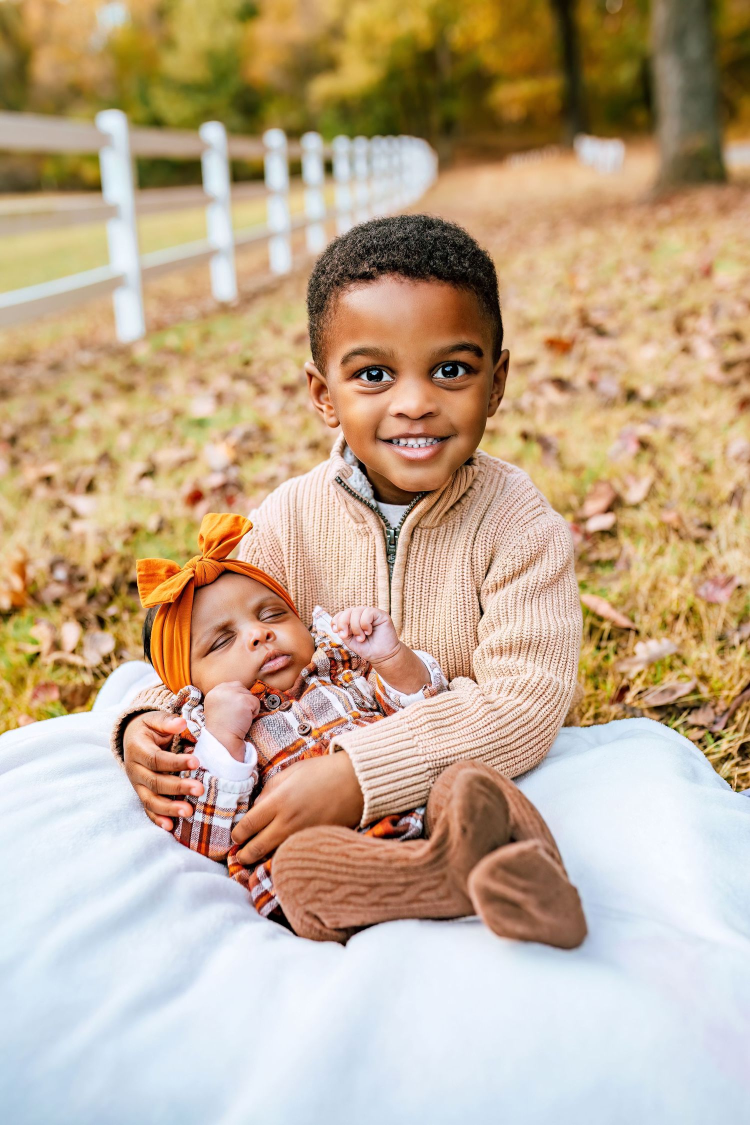 Children sitting on blanket by white fence during autumn photoshoot.