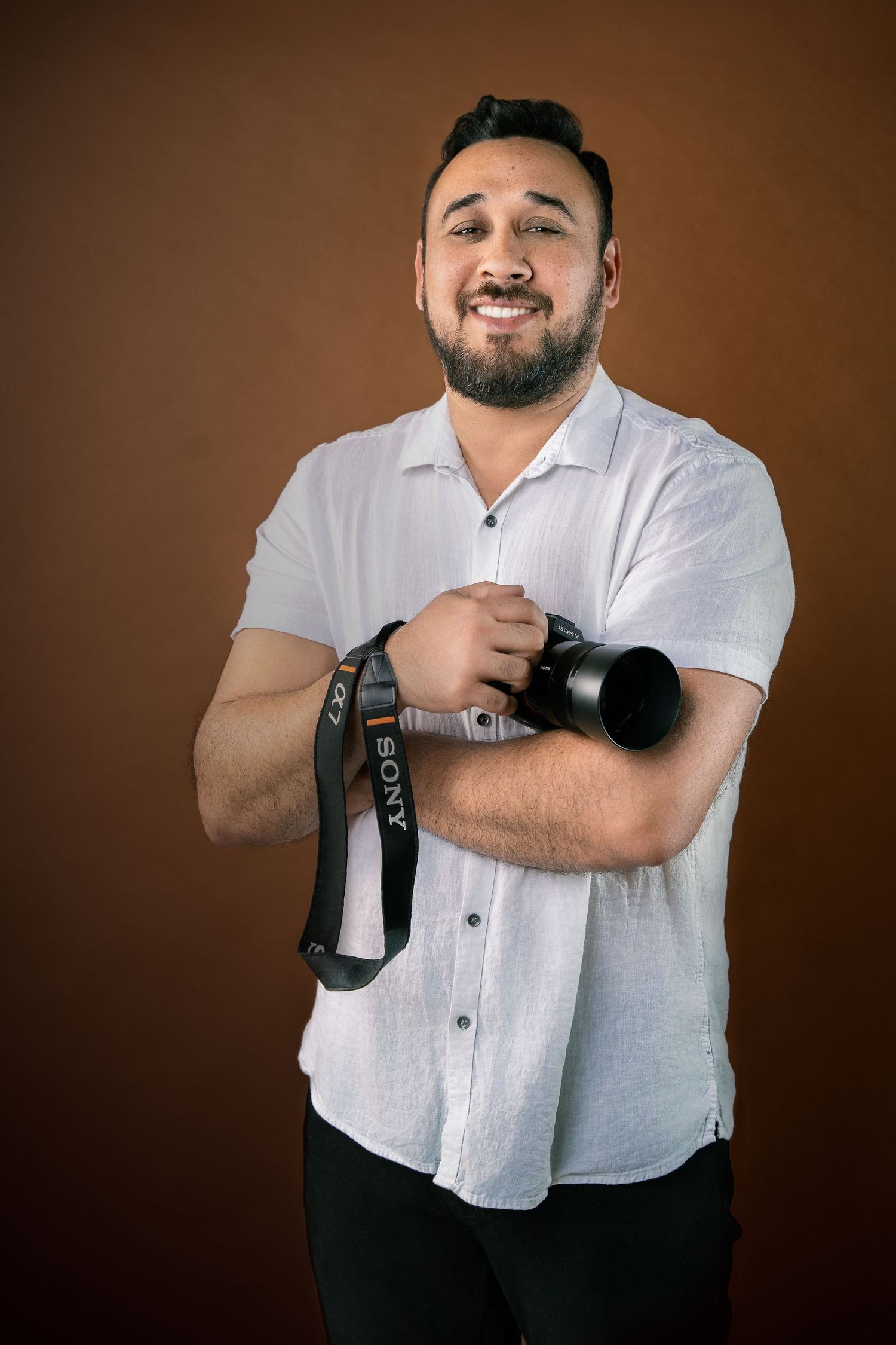 Photographer in white shirt smiles while holding professional camera against brown background.