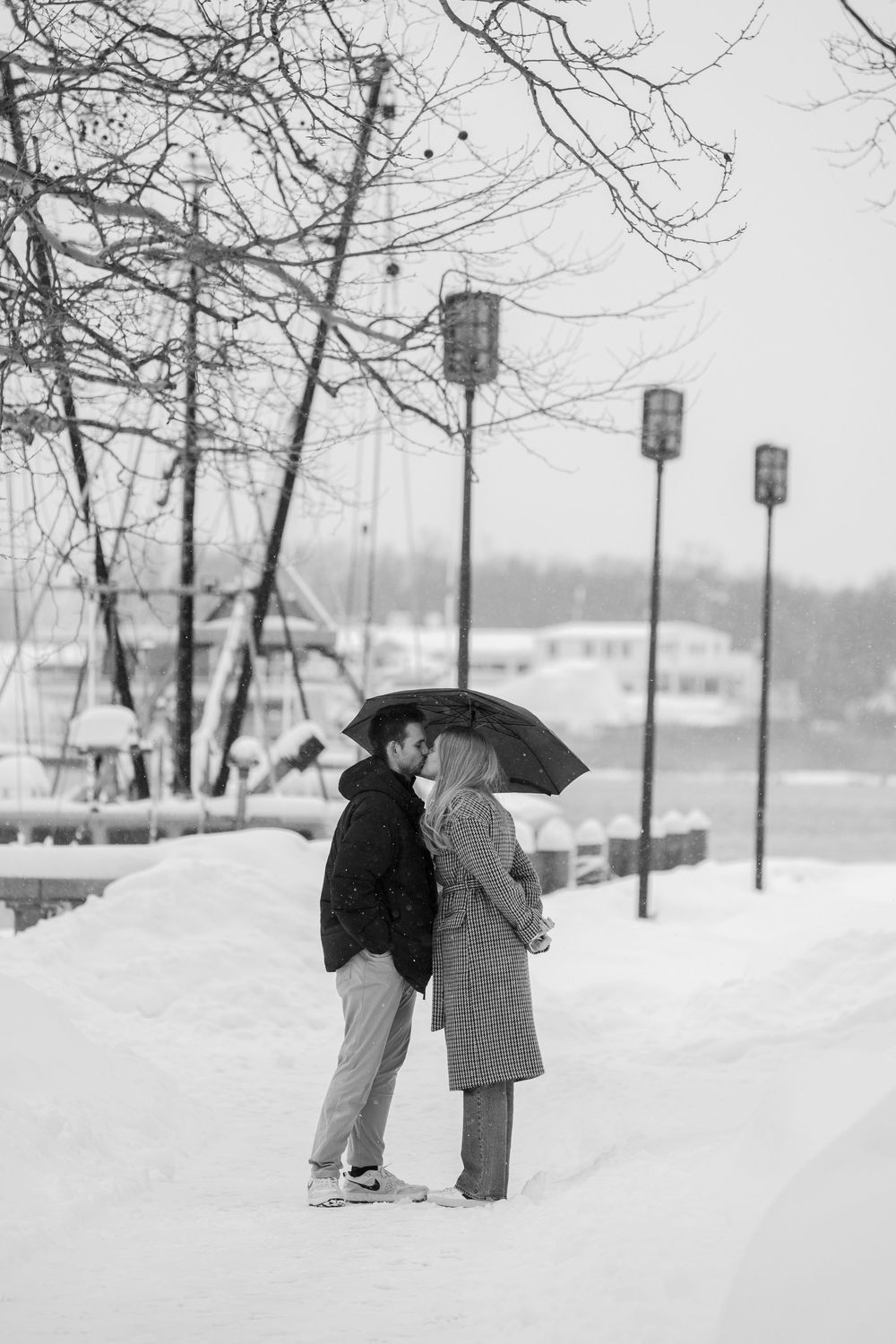 Two people in winter coats embrace on a snowy path surrounded by bare trees and street lamps.