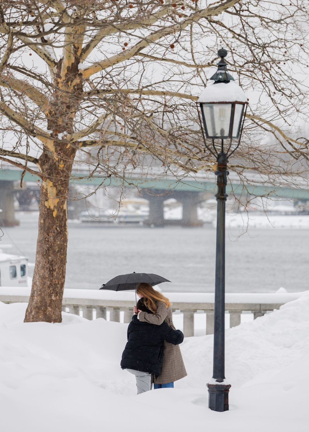 A person with an umbrella stands in snowy winter scene by lamppost near river with bridge in background.