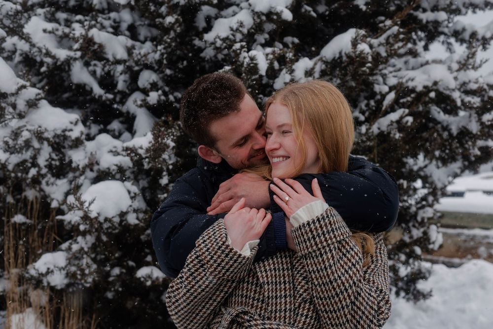 A couple shares intimate moments in a snowy winter setting with evergreen trees in the background.
