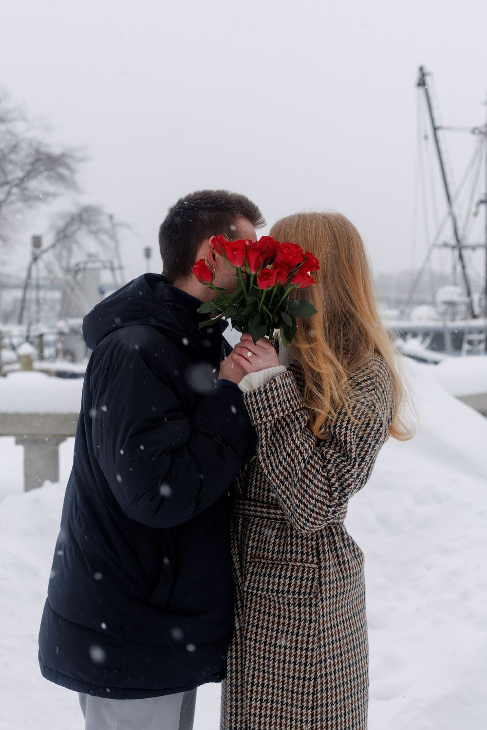 A couple shares a romantic moment with red roses in a snowy marina setting.