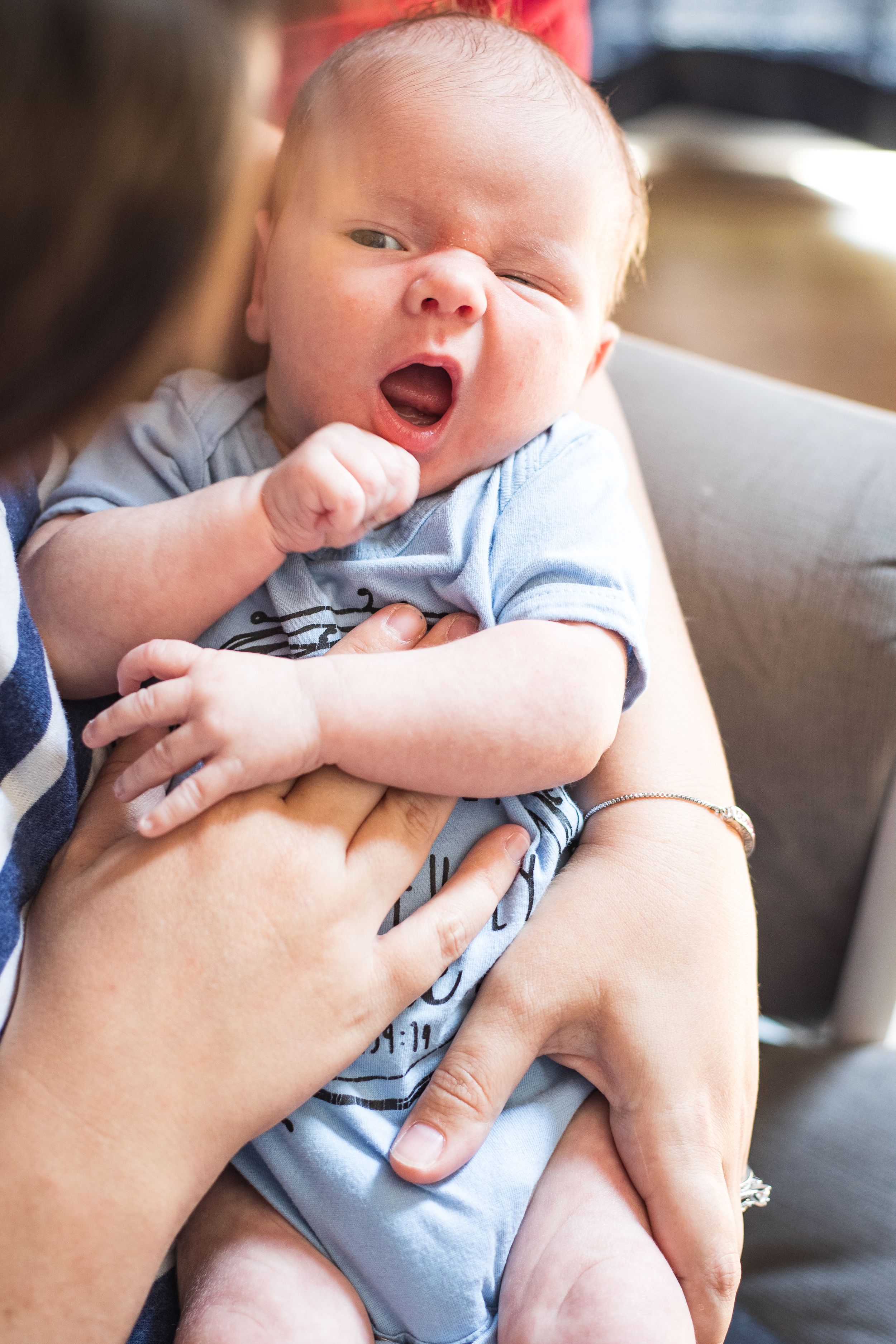 Close-up of a yawning newborn baby in a blue onesie cradled in mother's arms.