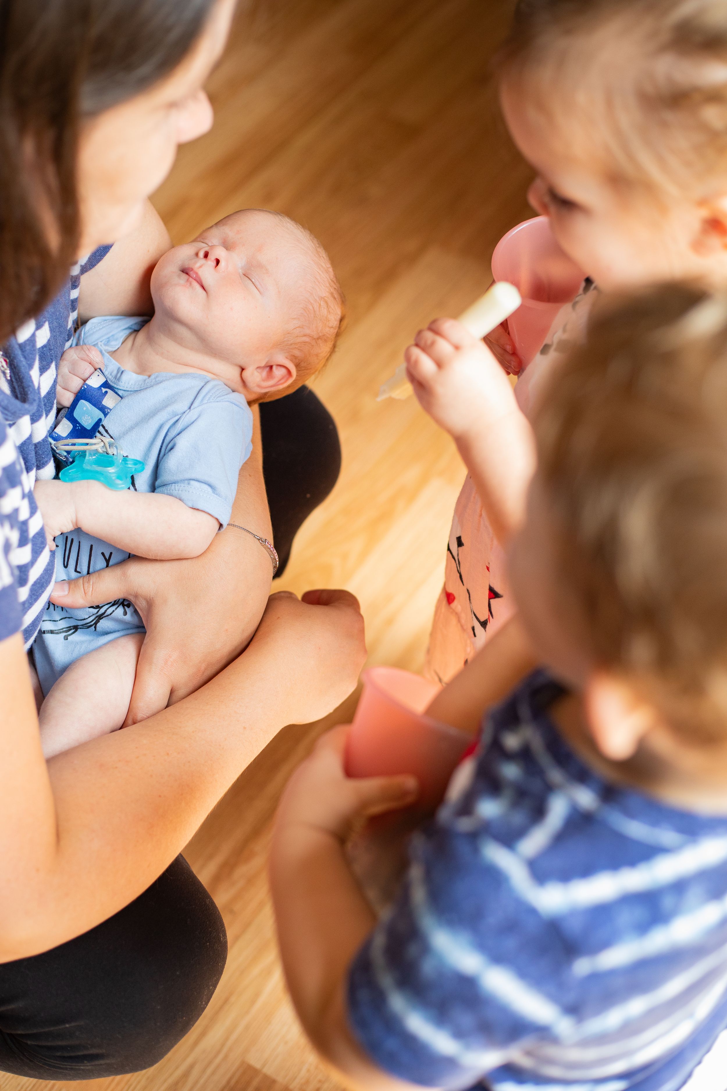 A woman holds a sleeping newborn in blue while children interact nearby on a wooden floor.
