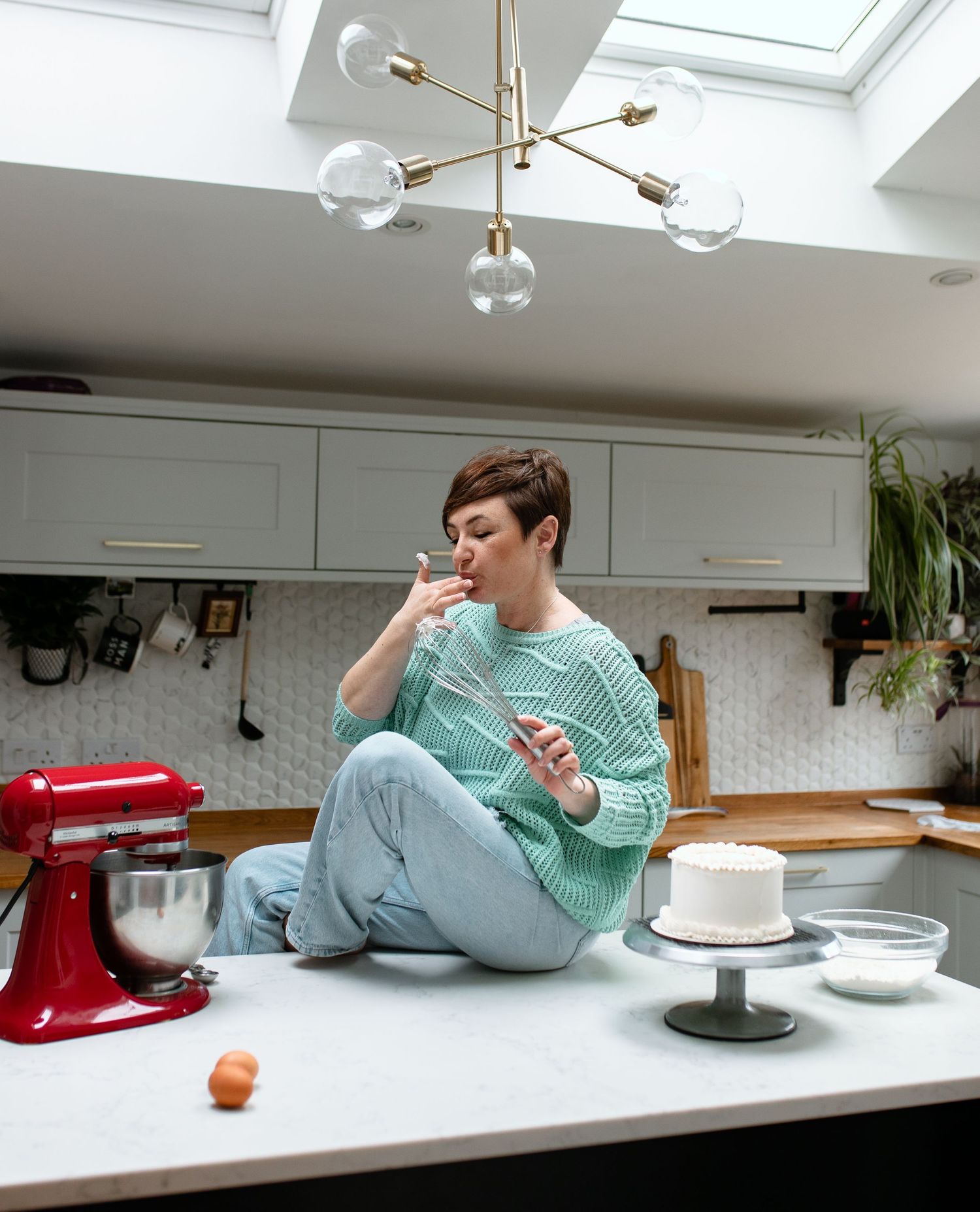 A sequence of images shows someone in a mint green sweater lying on a white countertop while interacting with a red mixer.