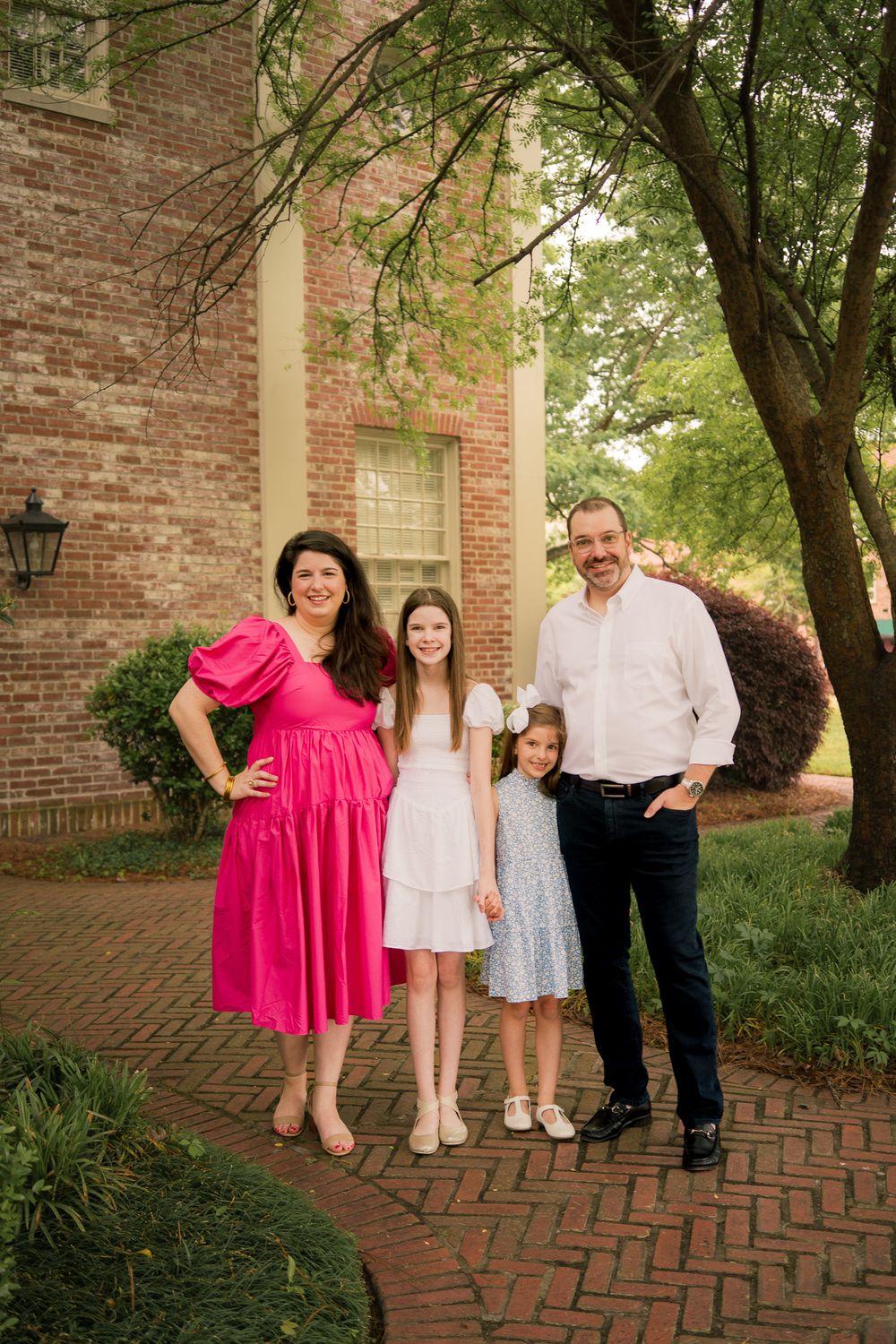 Family photo session on brick pathway with pink and white outfits near colonial style building.