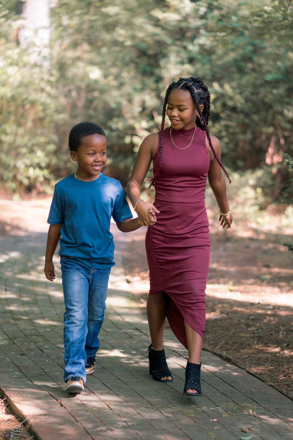 A pair walking together on a wooded path, one in a burgundy dress and one in blue jeans and t-shirt.