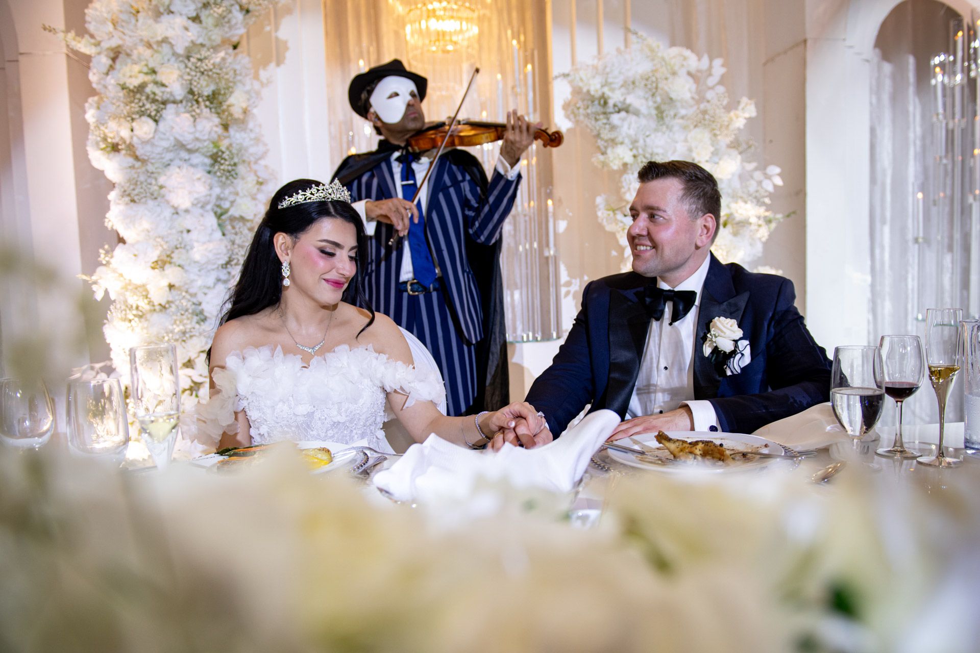 Bride and groom hold hands at sweetheart table while violinist performs at luxurious floral wedding reception.