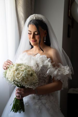 Radiant bride in tiara and veil holding white rose bouquet near window in soft natural light.