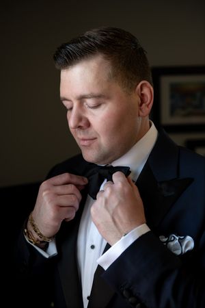 Groom in black tuxedo adjusting his bow tie while getting ready for wedding ceremony.