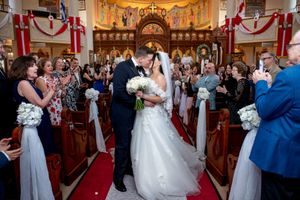 Bride and groom sharing first kiss at altar while guests applaud in decorated Orthodox church.